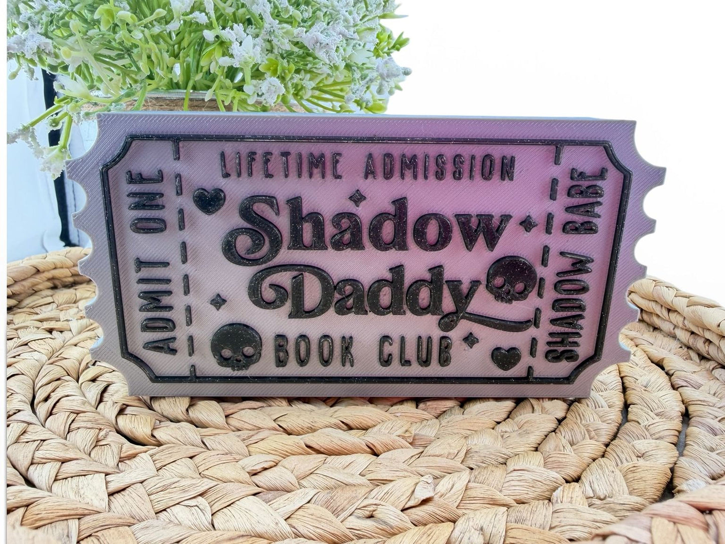 a ticket-shaped object with the text "Lifetime Admission Shadow Daddy Book Club" printed on it, placed on a woven surface.