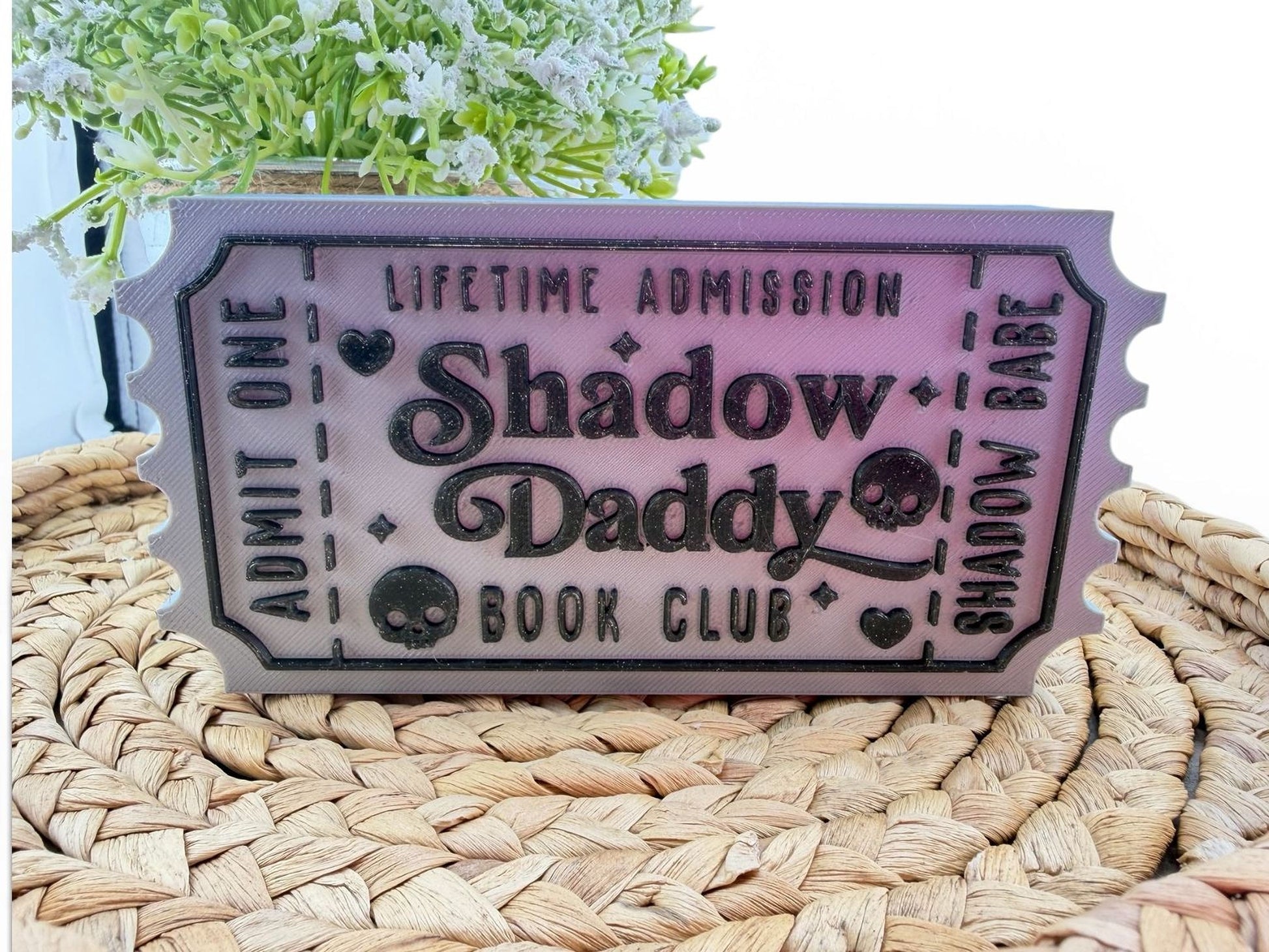 a ticket-shaped object with the text "Lifetime Admission Shadow Daddy Book Club" printed on it, placed on a woven surface.