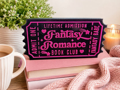 a pink and black ticket-shaped sign with the words "Fantasy Romance Book Club" written on it, placed on top of a stack of books and a white mug.