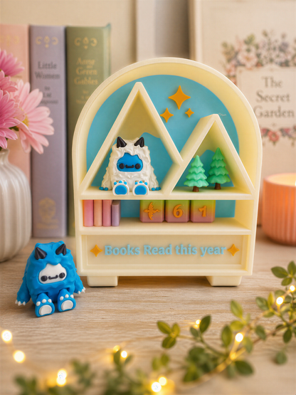 A whimsical bookshelf with a blue and yellow design, featuring a small blue monster figurine and a green monster figurine, along with a sign that says "Books Read this year". The bookshelf is surrounded by various books, a vase of flowers, and twinkling lights, creating a cozy and inviting atmosphere.