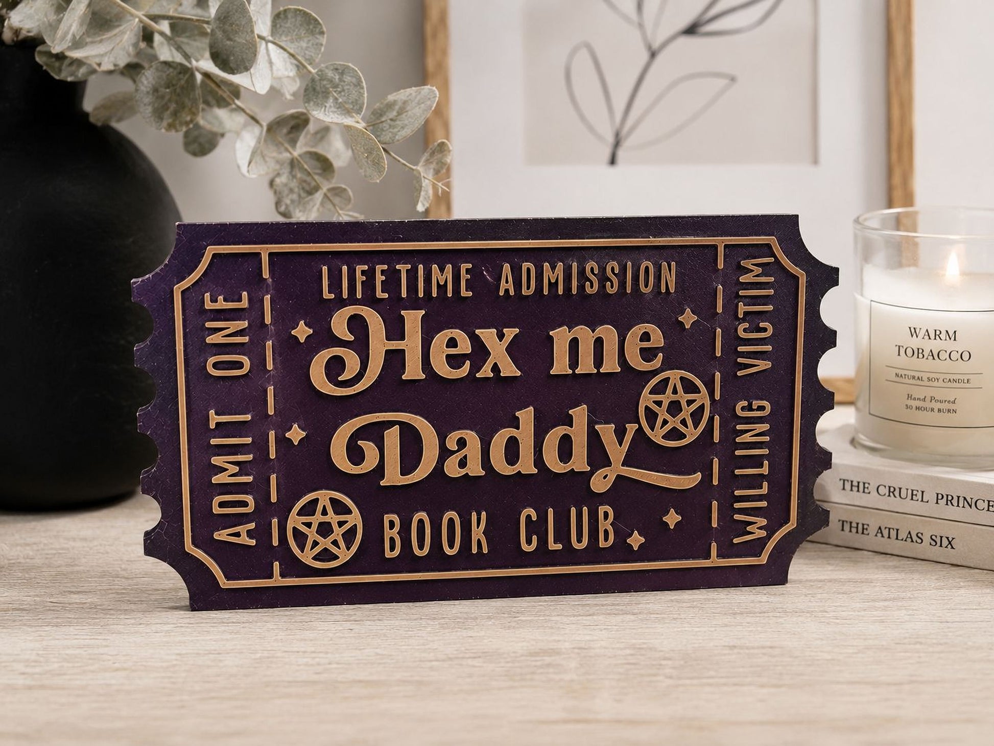 a purple ticket-shaped sign with the text "Hex me Daddy Book Club" on it, placed on a wooden surface. There is also a candle and some books in the background.