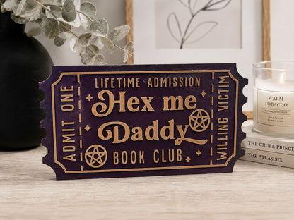 a purple ticket-shaped sign with the text "Hex me Daddy Book Club" on it, placed on a wooden surface. There is also a candle and some books in the background.