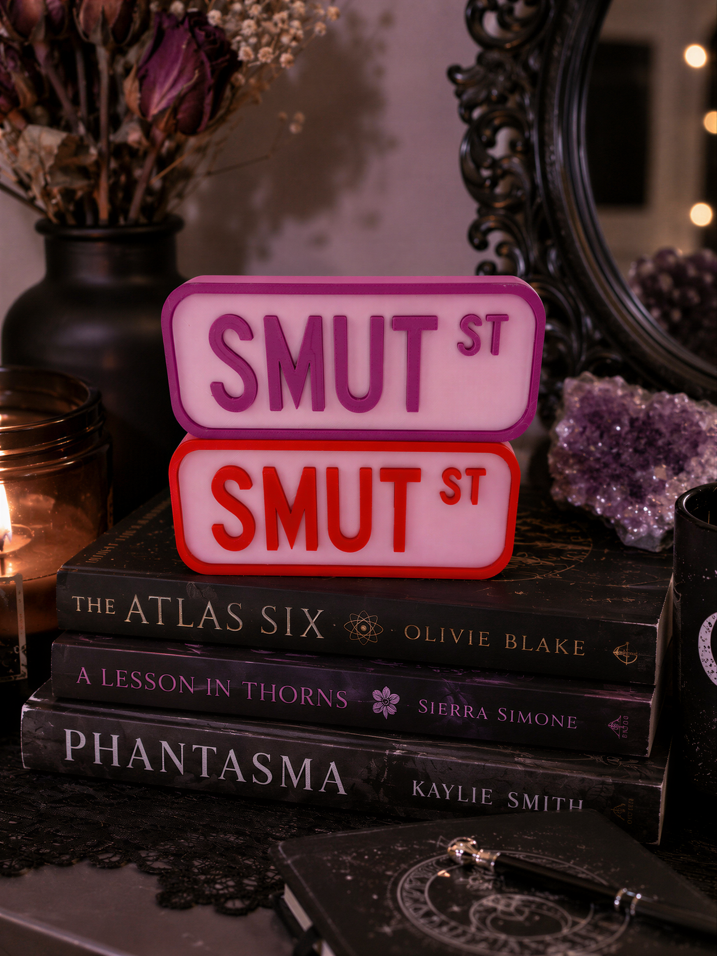 a stack of books, with a sign that says "SMUT ST" in pink and red letters. The books are titled "THE ATLAS SIX", "A LESSON IN THORNS", "PHANTASMA", and "KAYLIE SMITH". The background includes a vase with flowers and a mirror.