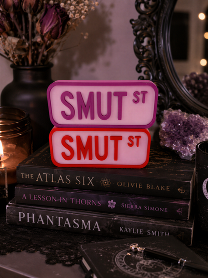 a stack of books, with a sign that says "SMUT ST" in pink and red letters. The books are titled "THE ATLAS SIX", "A LESSON IN THORNS", "PHANTASMA", and "KAYLIE SMITH". The background includes a vase with flowers and a mirror.