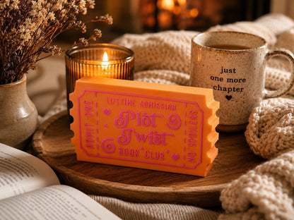 a cozy scene with a book, a mug, and a ticket stub on a wooden tray. The book is open, and the mug has a quote on it. The ticket stub is orange and pink, and it appears to be a ticket stub for a book club.