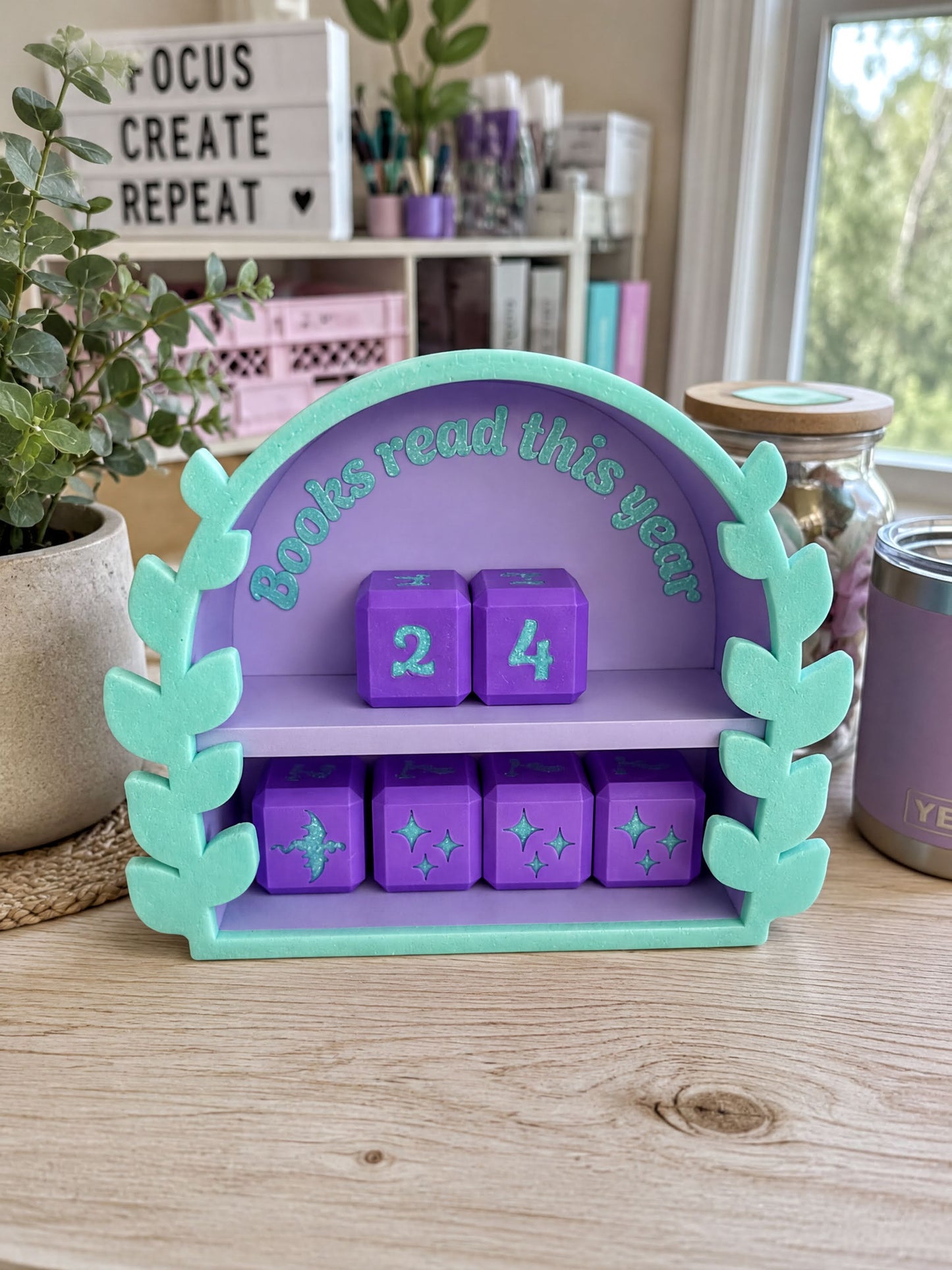 A colorful bookshelf with a purple and green design, displaying a series of purple cubes with numbers and stars, and a sign that says "Books Read this year". The shelf is placed on a wooden surface, with various items such as a plant, a jar, and a book visible in the background.