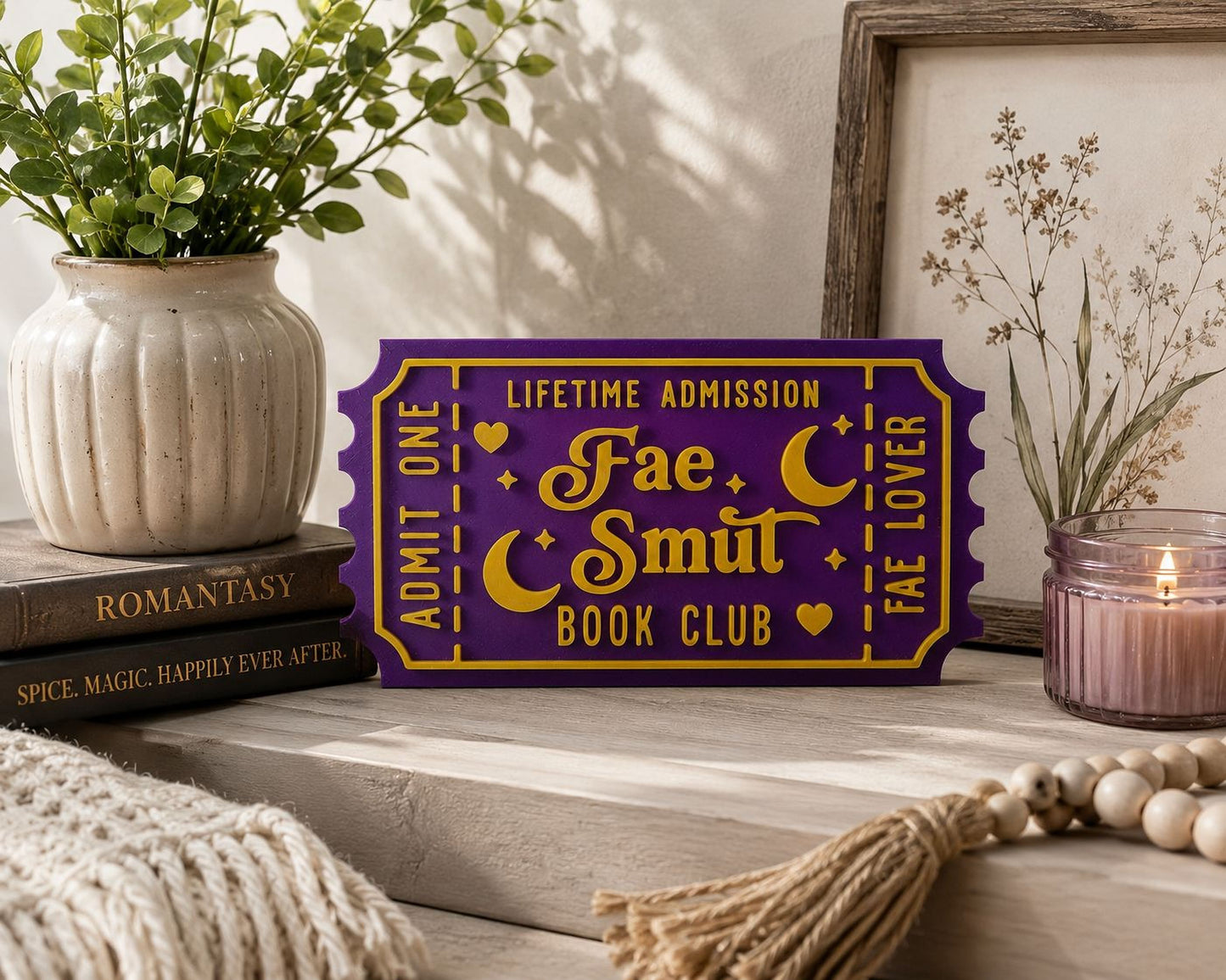 a purple ticket-shaped sign with gold lettering, placed on a wooden surface next to a candle, a plant, and some books.
