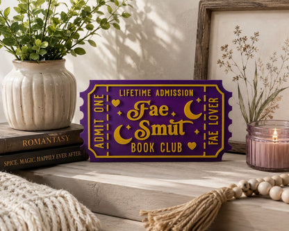 a purple ticket-shaped sign with gold lettering, placed on a wooden surface next to a candle, a plant, and some books.
