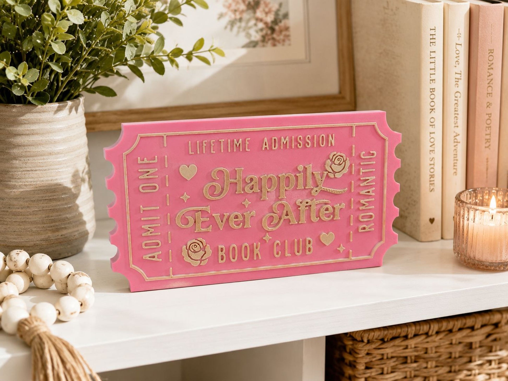 a pink ticket-shaped sign with the words "Happy Ever After" on it, placed on a white shelf next to some books and a candle.