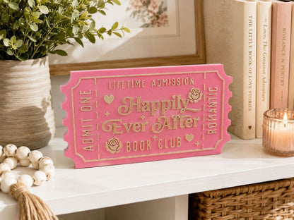 a pink ticket-shaped sign with the words "Happy Ever After" on it, placed on a white shelf next to some books and a candle.