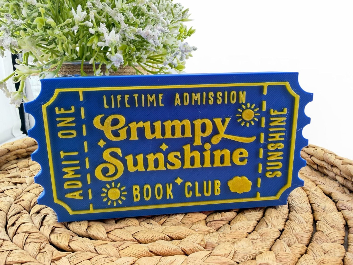 A blue and yellow ticket-shaped sign with the text "Lifetime Admission Grumpy Sunshine Book Club" on it, placed on a woven mat.