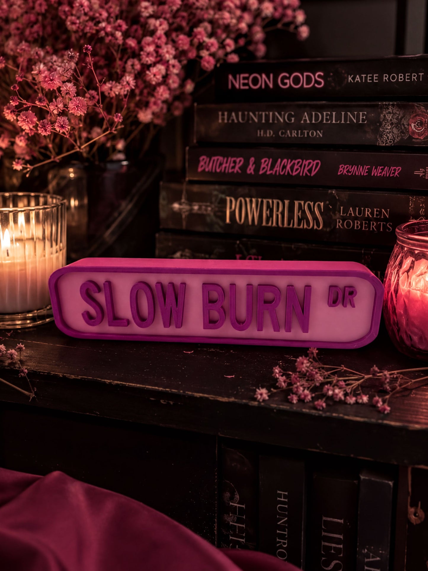 a collection of books on a shelf, with a pink sign that reads "SLOW BURN DR" placed in front of them. The books include titles like "Neon Gods", "Haunting Adeline", "Butcher & Blackbird", "Powerless", and "Lauren Roberts". The scene is set against a backdrop of pink flowers, creating a cozy and inviting atmosphere.
