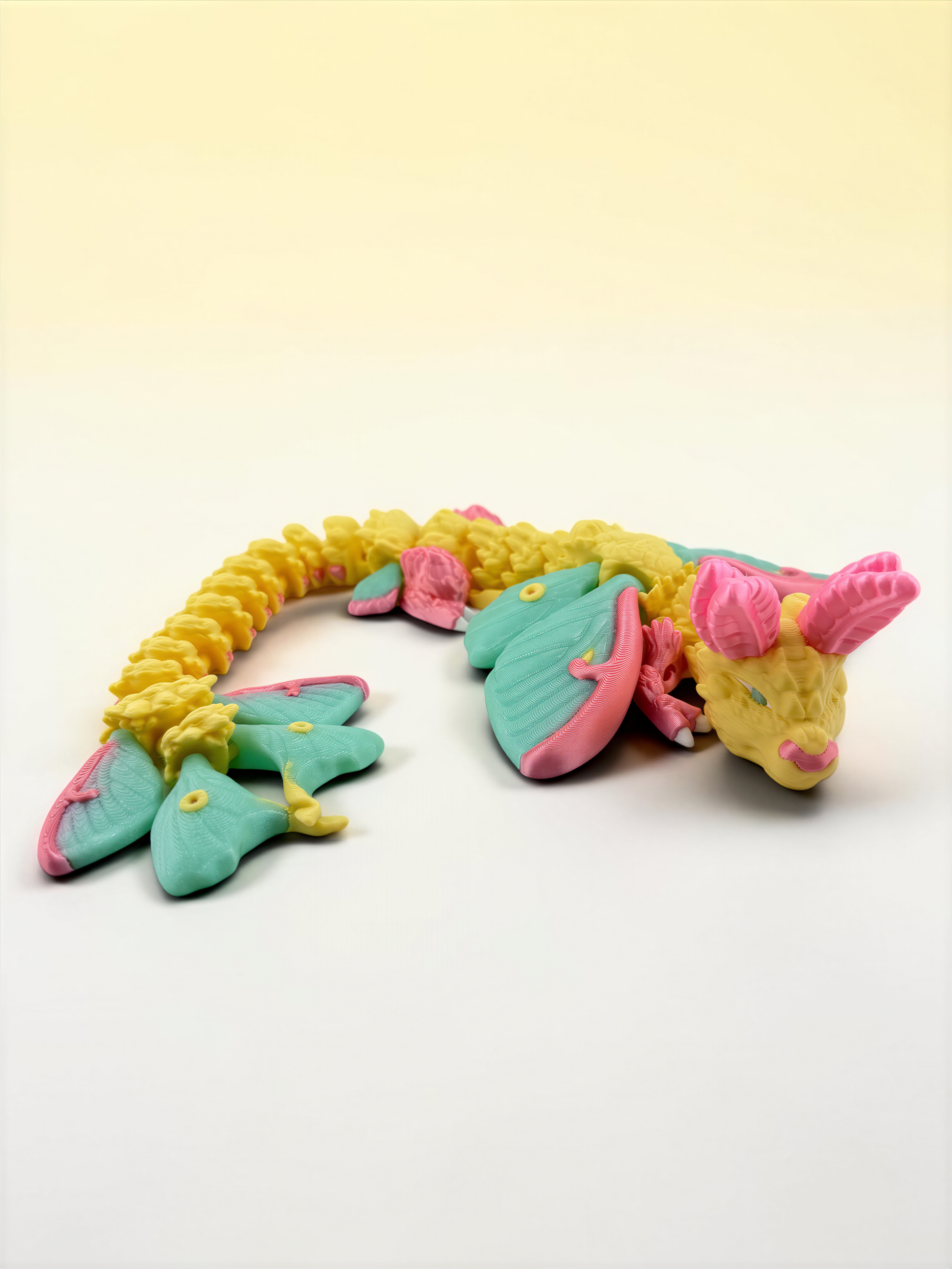 A colorful toy dragon made of plastic or clay, with a yellow body, green wings, and pink accents, is lying on a plain white background.