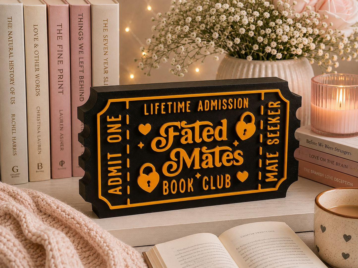 a black and gold ticket-shaped sign with the text "Lifetime Admission Fated Mates Book Club" on it, placed on a table surrounded by books, a vase of flowers, and a candle.