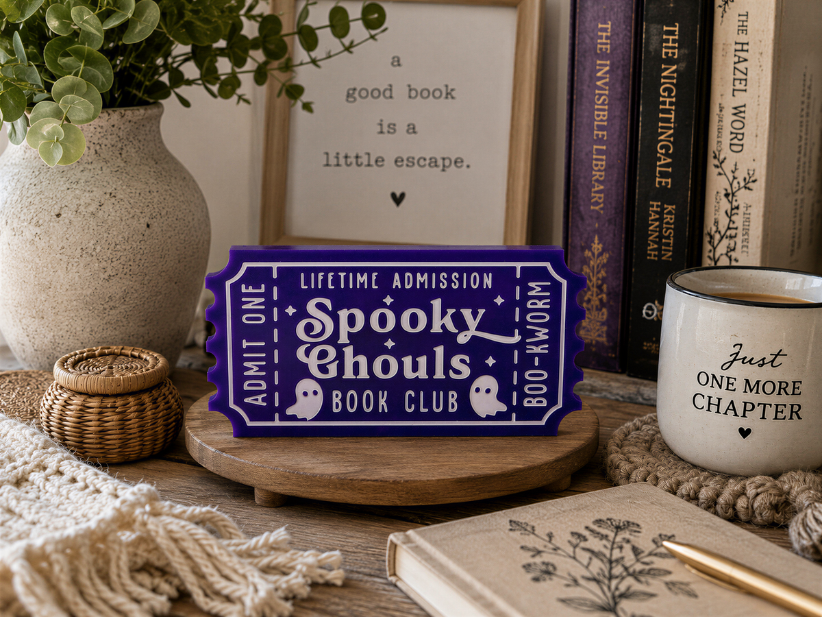 a purple ticket-shaped sign for a book club, surrounded by various items such as a mug, a plant, and books.
