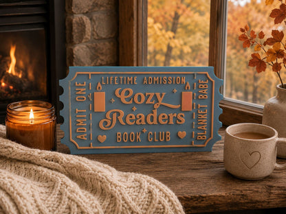 A cozy reading sign, a candle, a mug, and a vase with flowers are arranged on a wooden surface, with a fireplace visible in the background.