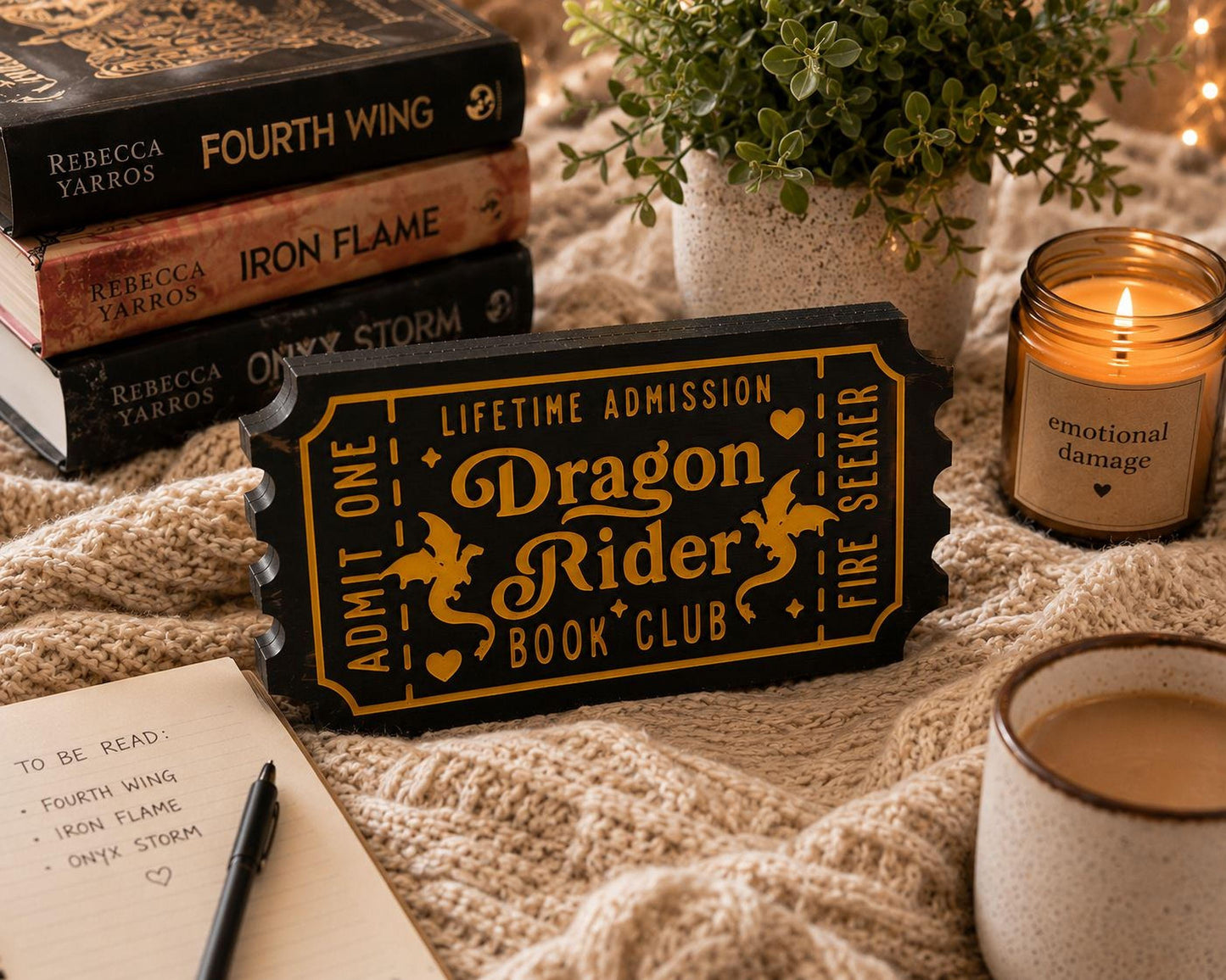 a cozy scene with a stack of books, a potted plant, a candle, and a notebook. The books are titled "Fourth Wing", "Iron Flame", and "Only Storm", suggesting a fantasy or adventure theme. The potted plant adds a touch of greenery, while the candle provides a warm ambiance. The notebook and pen indicate that someone might be planning to read or write about the books.