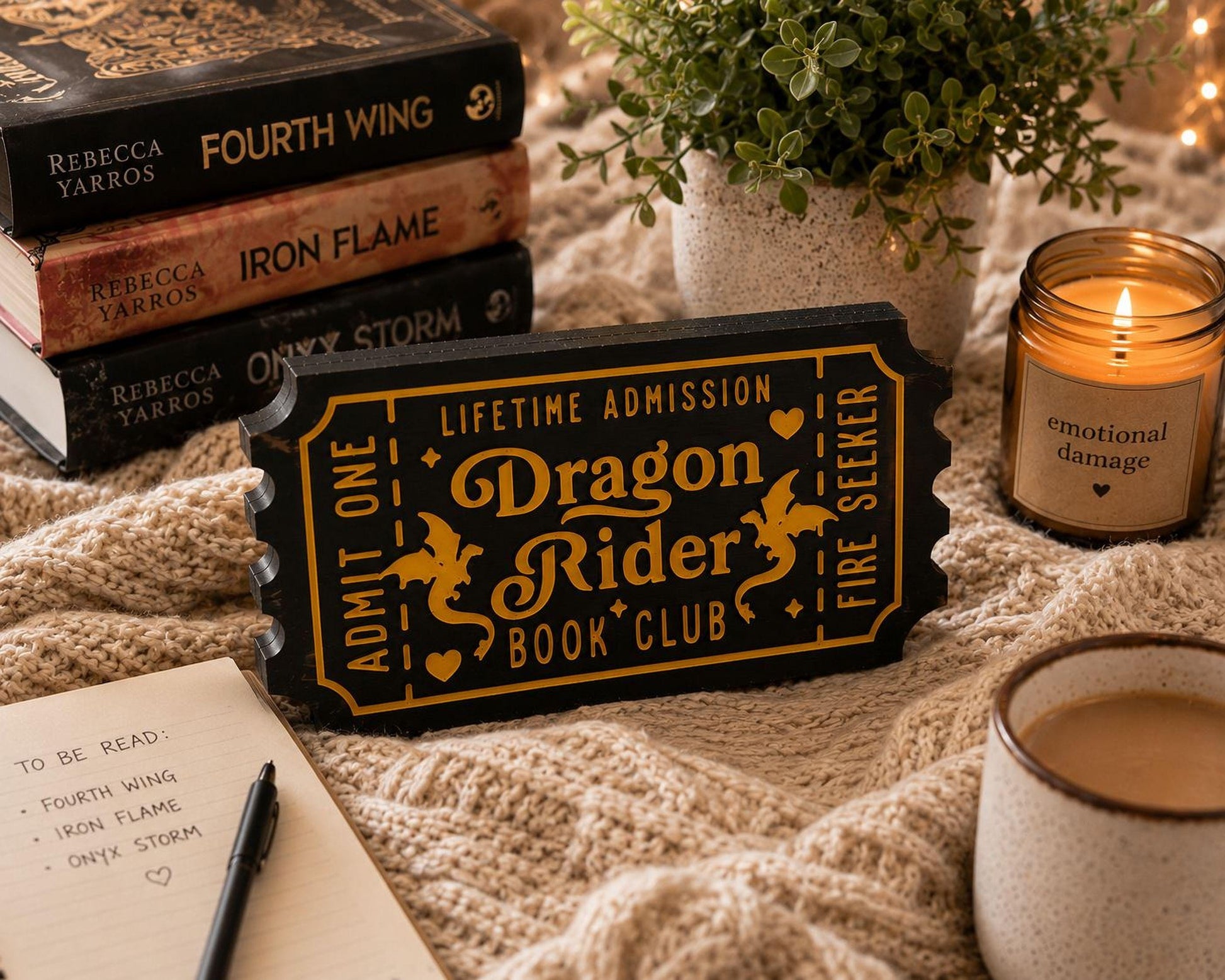 a cozy scene with a stack of books, a potted plant, a candle, and a notebook. The books are titled "Fourth Wing", "Iron Flame", and "Only Storm", suggesting a fantasy or adventure theme. The potted plant adds a touch of greenery, while the candle provides a warm ambiance. The notebook and pen indicate that someone might be planning to read or write about the books.