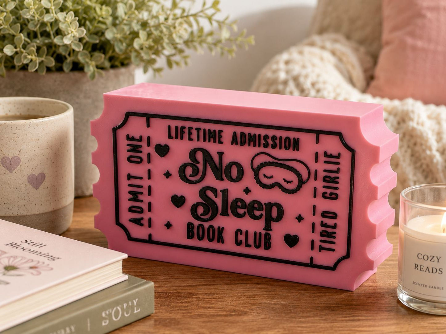 a pink ticket-shaped object with text and a sleep mask design, placed on a wooden surface next to some books and a candle.