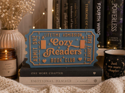 a cozy reading nook with a blue ticket-shaped sign that reads "Lifetime Admission Cozy Readers Book Club" surrounded by books, a candle, and a mug.