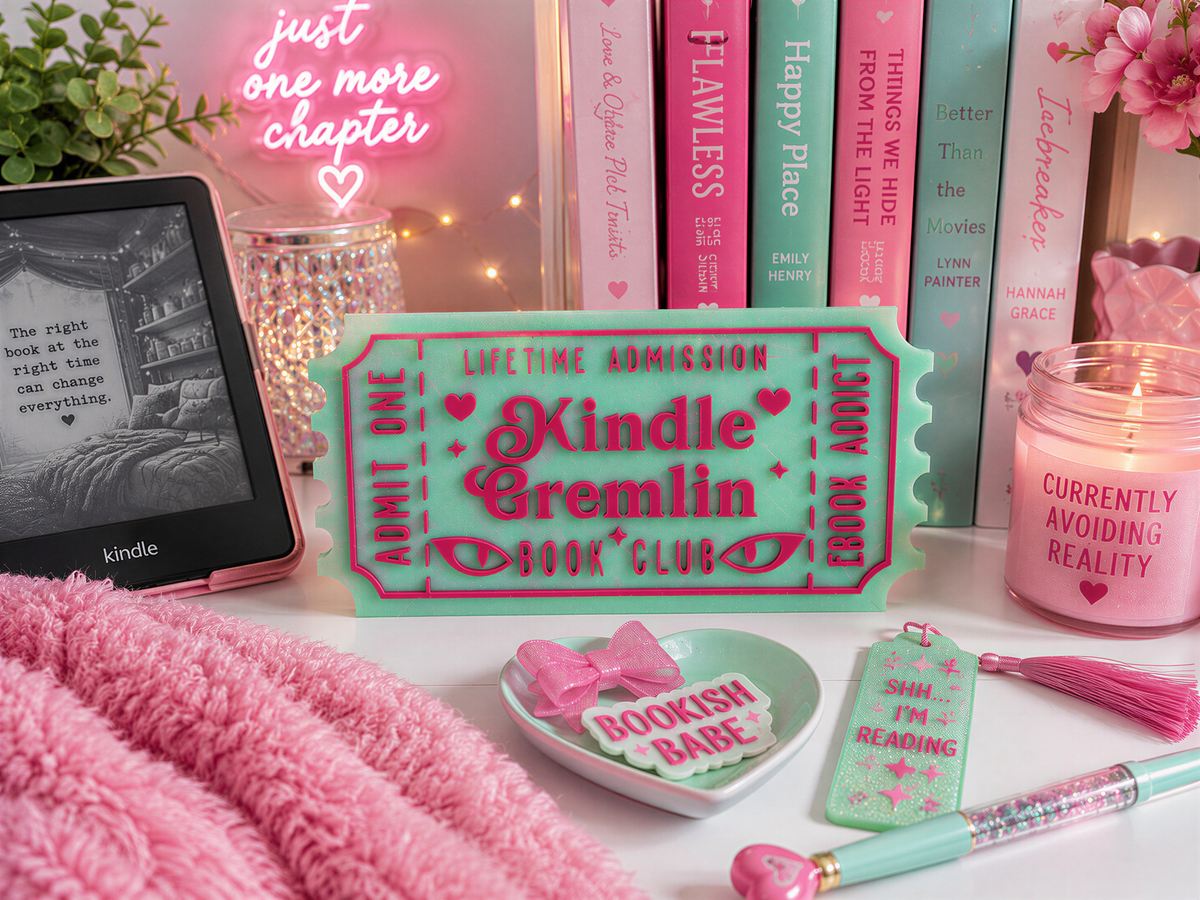 a collection of pink and green items, including a Kindle e-reader, a pink candle, a pink tassel, a pink bow, and a pink book club sign, all arranged on a white surface.