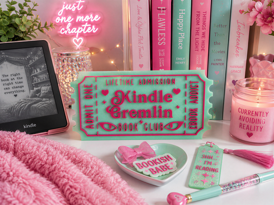a collection of pink and green items, including a Kindle e-reader, a pink candle, a pink tassel, a pink bow, and a pink book club sign, all arranged on a white surface.