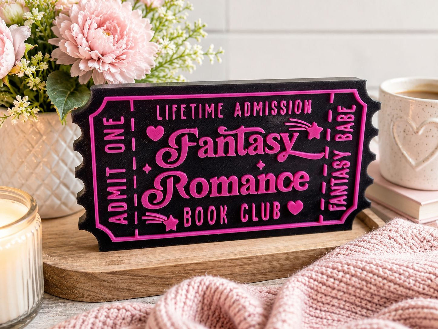a pink and black ticket-shaped sign that says "Lifetime Admission Fantasy Romance Book Club", surrounded by pink flowers, a candle, and a mug.
