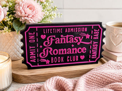a pink and black ticket-shaped sign that says "Lifetime Admission Fantasy Romance Book Club", surrounded by pink flowers, a candle, and a mug.