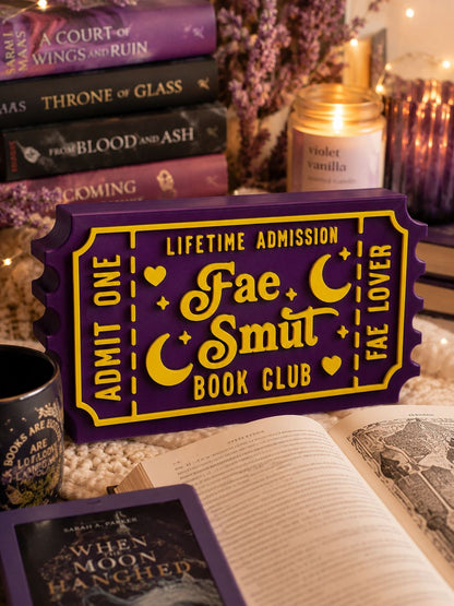 a purple ticket-shaped object with text, surrounded by various books, a candle, and a mug, creating a cozy and inviting atmosphere.