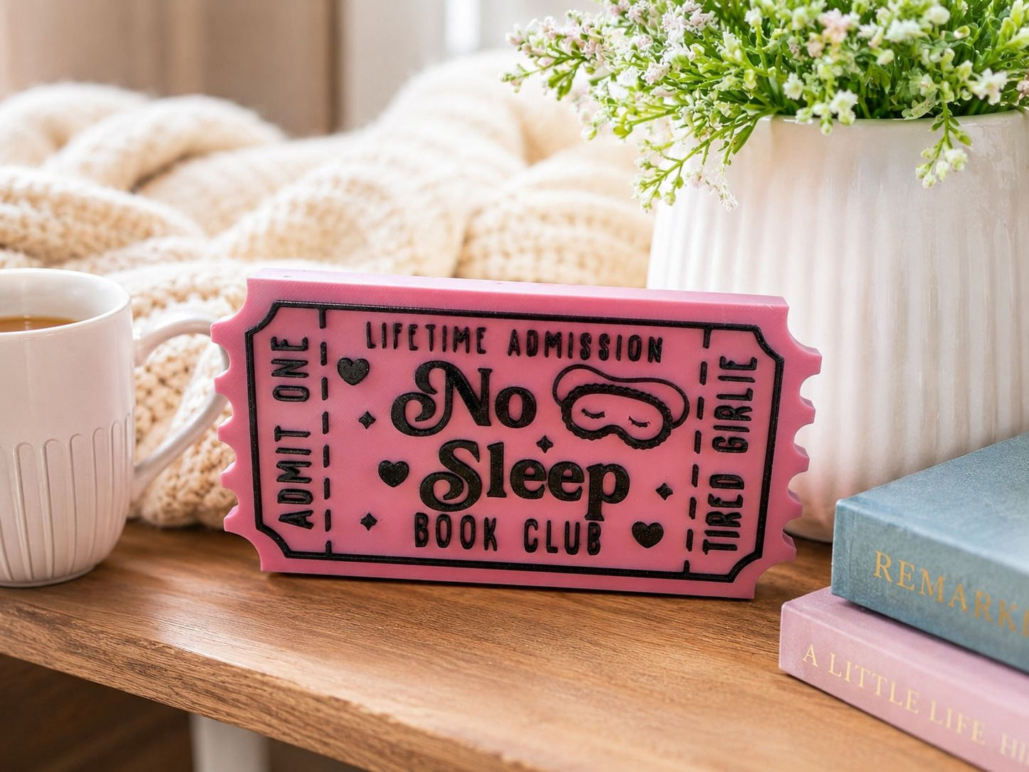 a pink ticket-shaped object with the text "No Sleep Book Club" on it, placed on a wooden surface next to a white mug, a white vase with flowers, and a stack of books.