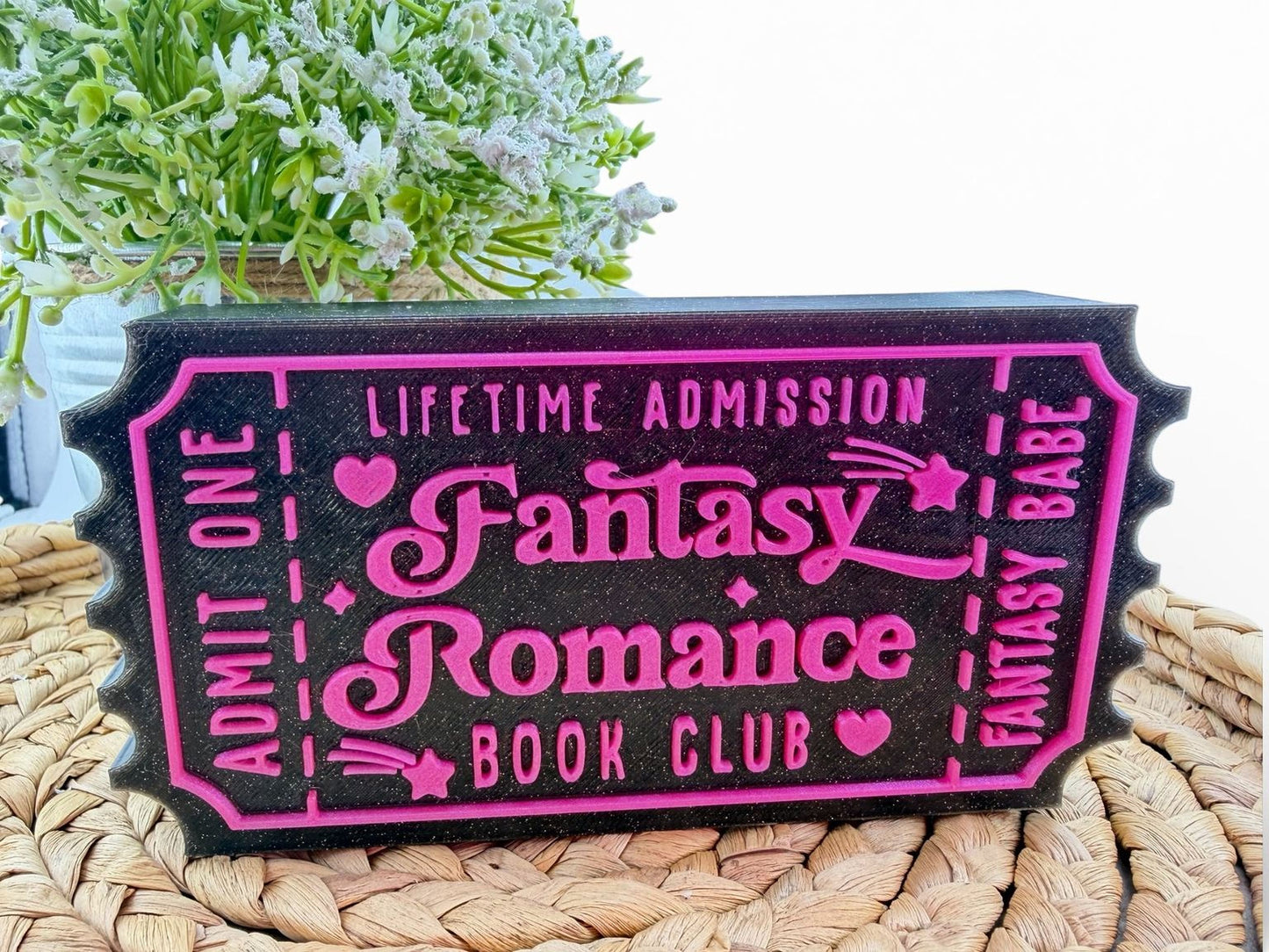 A black and pink fantasy romance book club ticket-shaped object with the text "Lifetime Admission Fantasy Romance Book Club" printed on it.