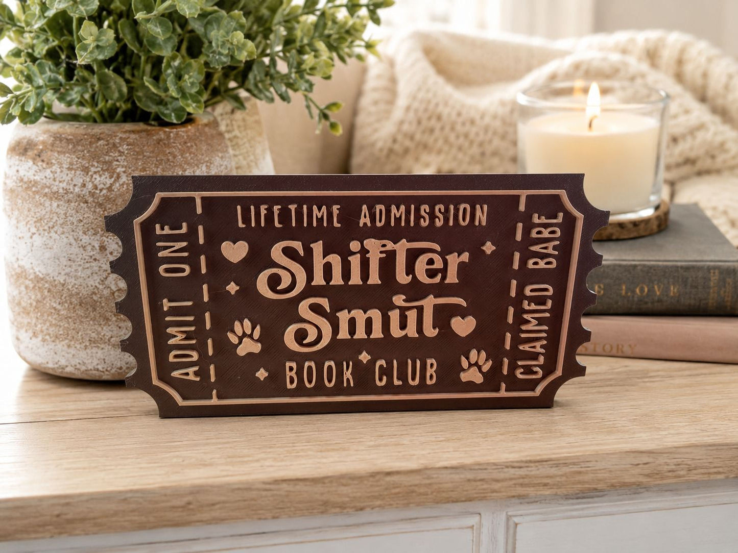 a brown ticket-shaped sign with the text "Shiftert Smut Book Club" on it, placed on a wooden surface next to a potted plant and a lit candle.