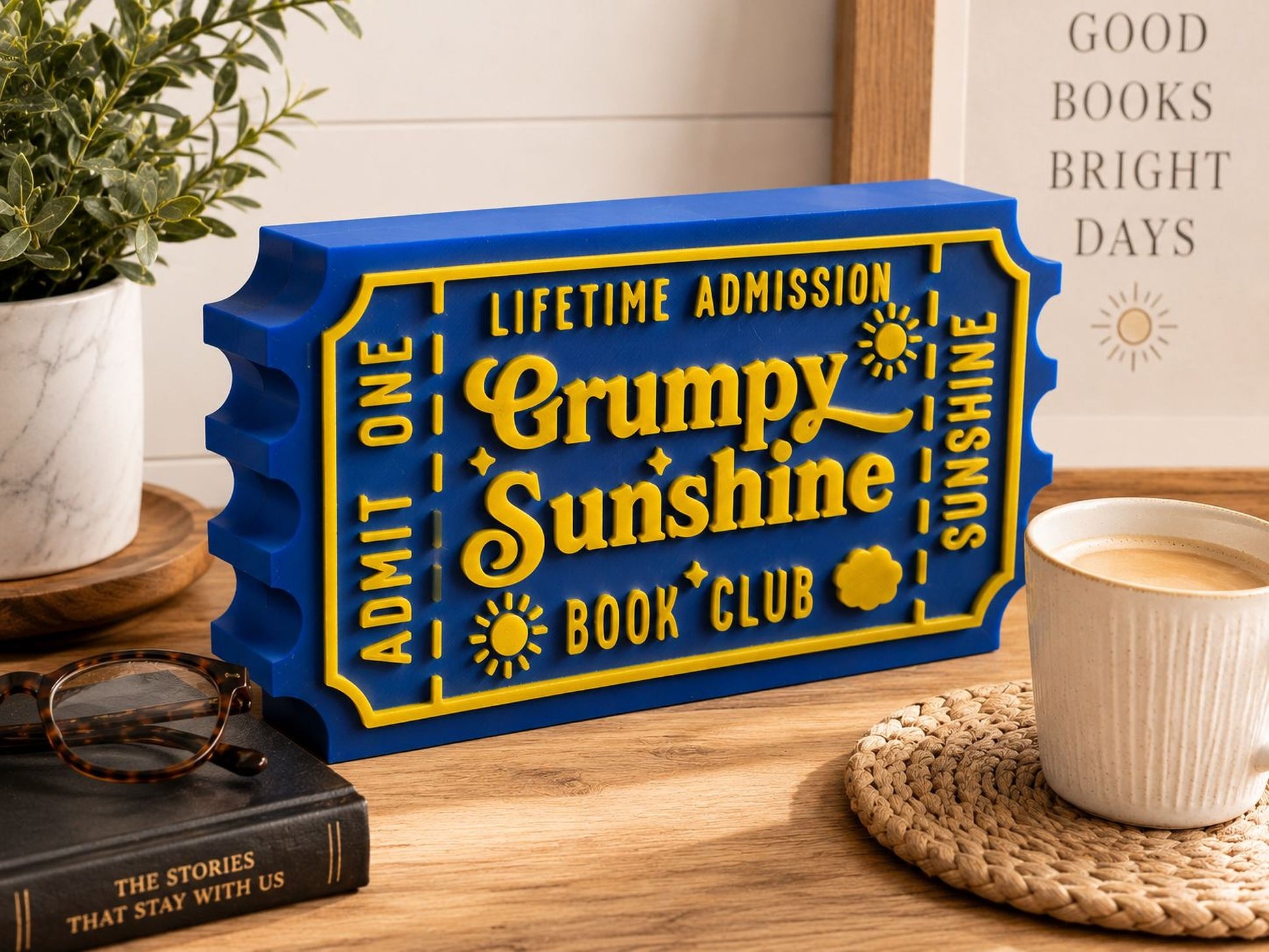 a blue and yellow ticket-shaped object with the text "Lifetime Admission Grumpy Sunshine Book Club" on it, placed on a wooden surface next to a mug of coffee and a book.