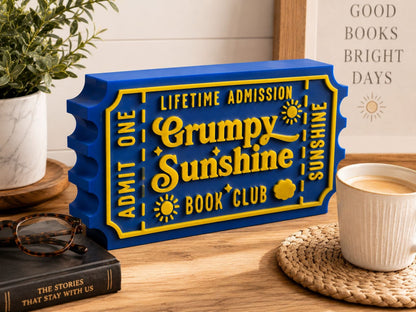 a blue and yellow ticket-shaped object with the text "Lifetime Admission Grumpy Sunshine Book Club" on it, placed on a wooden surface next to a mug of coffee and a book.