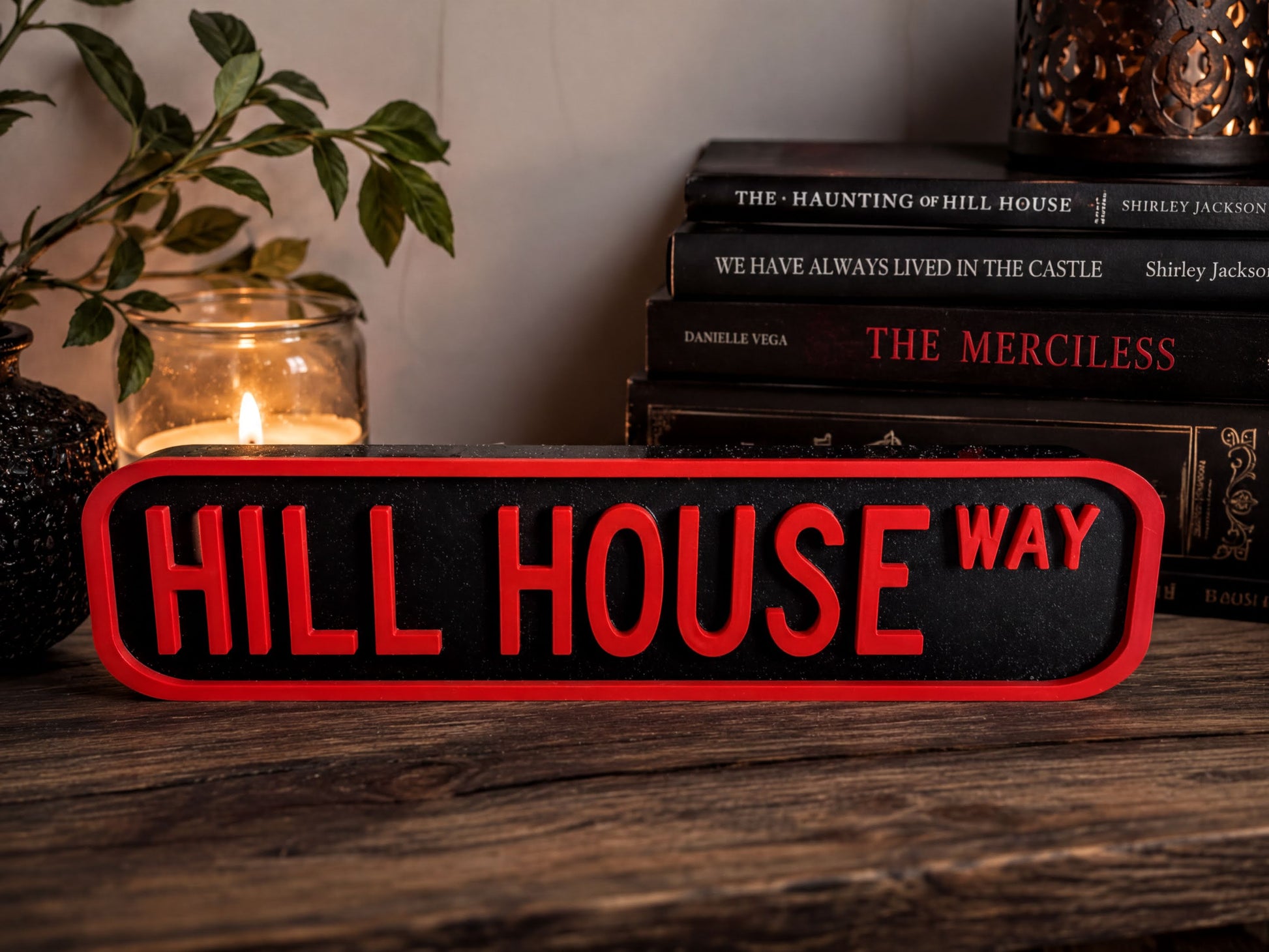 a sign that says "HILL HOUSE WAY" in bold red letters, placed on a wooden surface next to a stack of books and a lit candle.