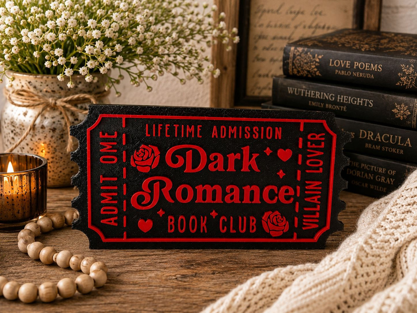 a red ticket-shaped sign with the words "Lifetime Admission" and "Dark Romance Book Club" written on it, placed on a wooden surface. There are also several books, a candle, and some flowers in the background.