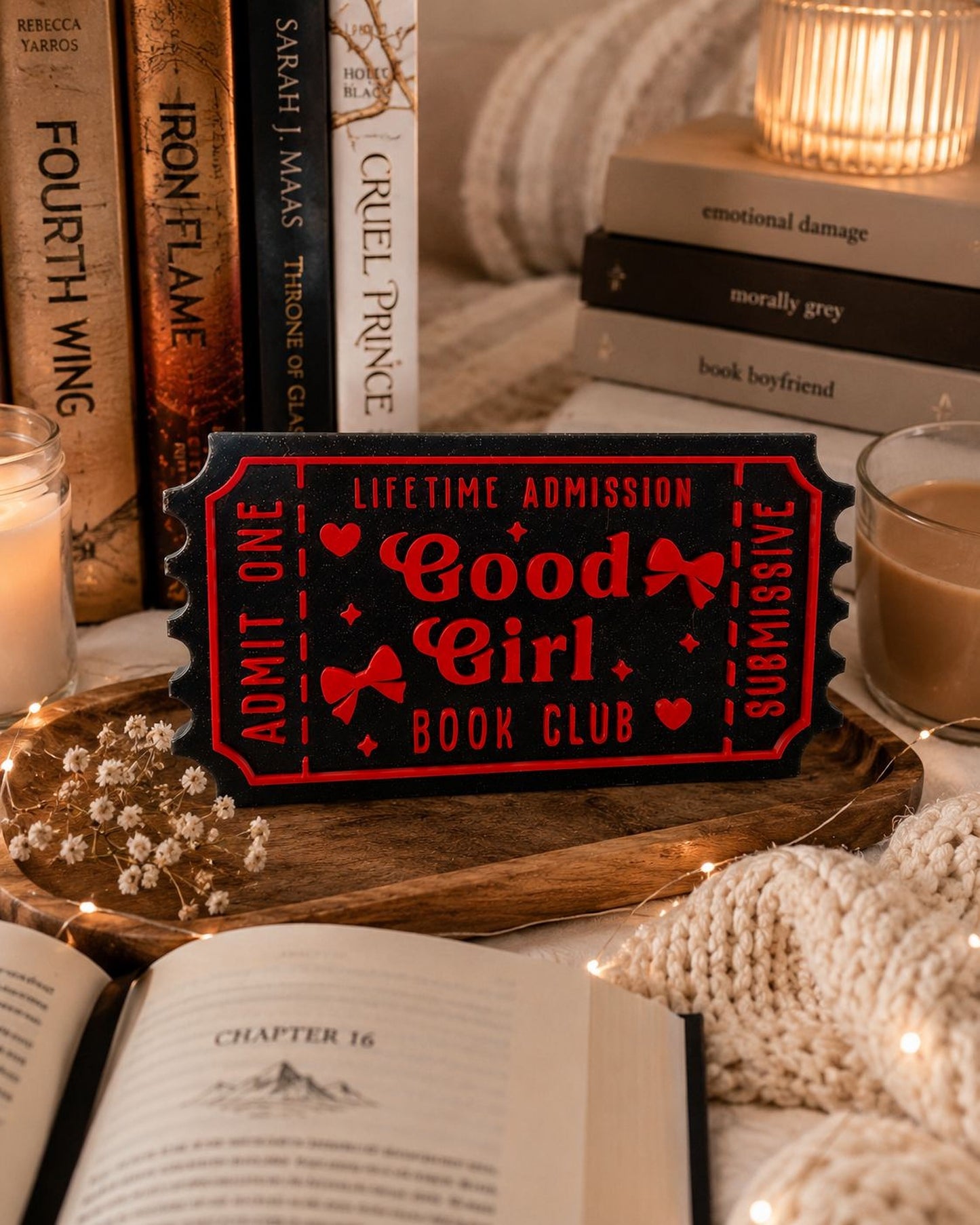a book club sign with the title "Good Girl Book Club" displayed prominently in the center. The sign is surrounded by various books, a candle, and a cozy blanket, creating a warm and inviting atmosphere.