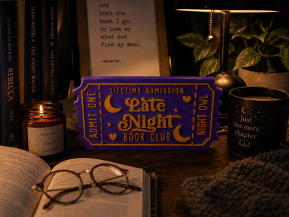 a cozy scene with a purple ticket for a book club event, a candle, a mug, a pair of glasses, and a book on a table.