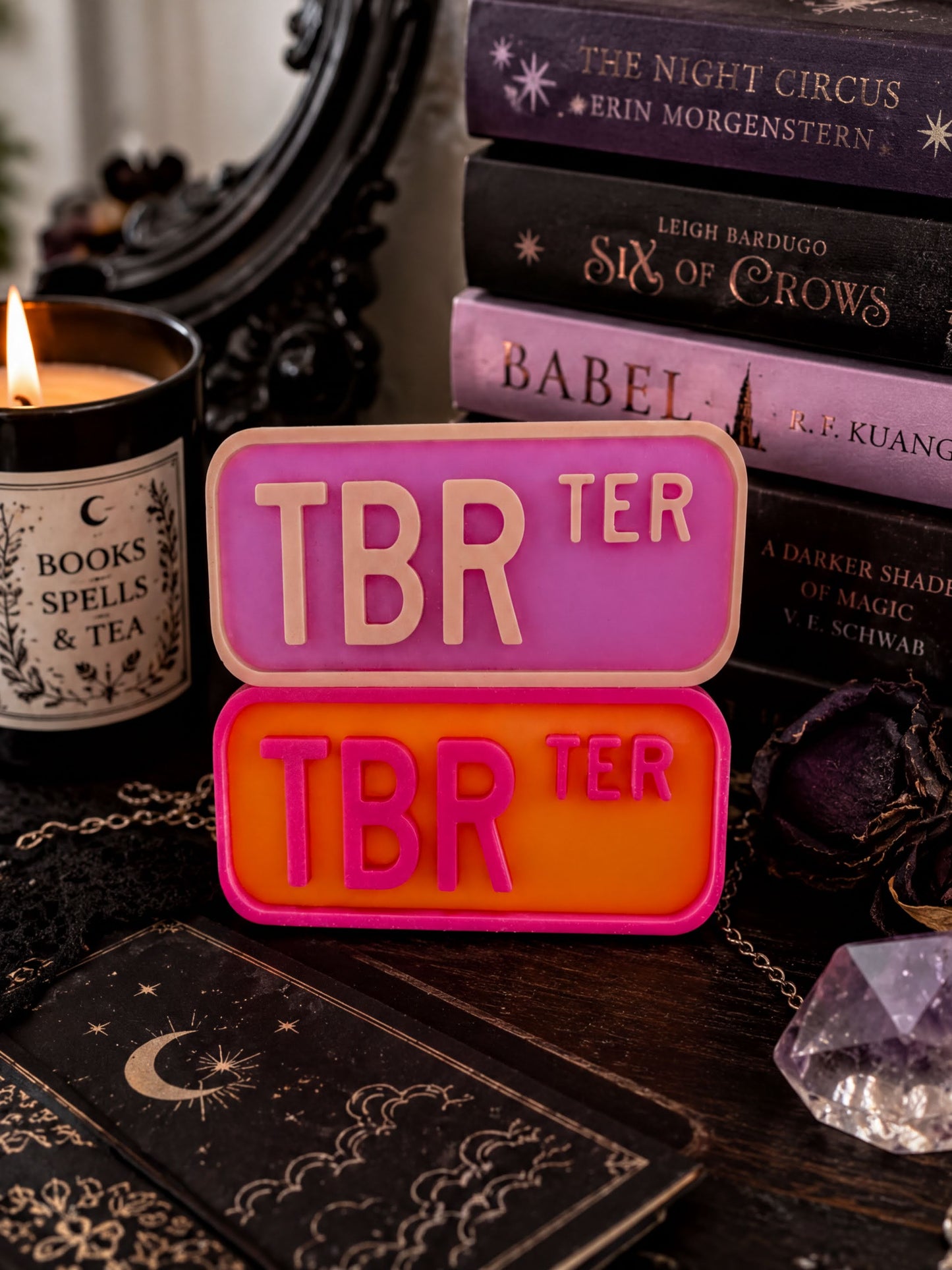 a collection of books, a candle, and a crystal on a table, with a pink and orange sign that says "TBR".