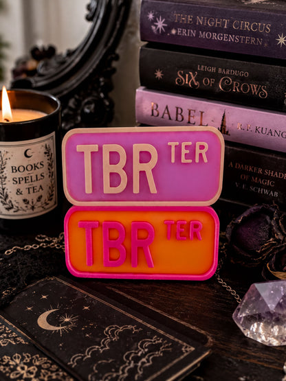 a collection of books, a candle, and a crystal on a table, with a pink and orange sign that says "TBR".