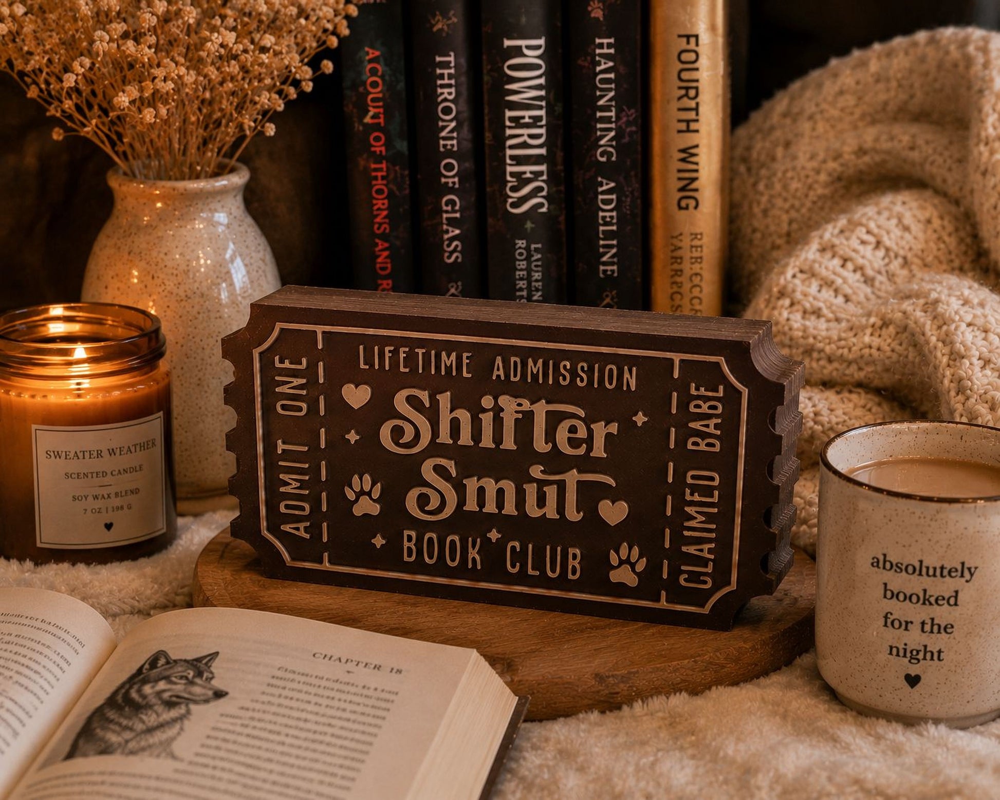 a cozy scene with a book titled "Shitter Smut" on a wooden stand, surrounded by various items such as a candle, a mug, and a book.