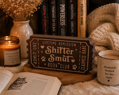 a cozy scene with a book titled "Shitter Smut" on a wooden stand, surrounded by various items such as a candle, a mug, and a book.