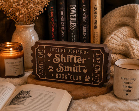 a cozy scene with a book titled "Shitter Smut" on a wooden stand, surrounded by various items such as a candle, a mug, and a book.
