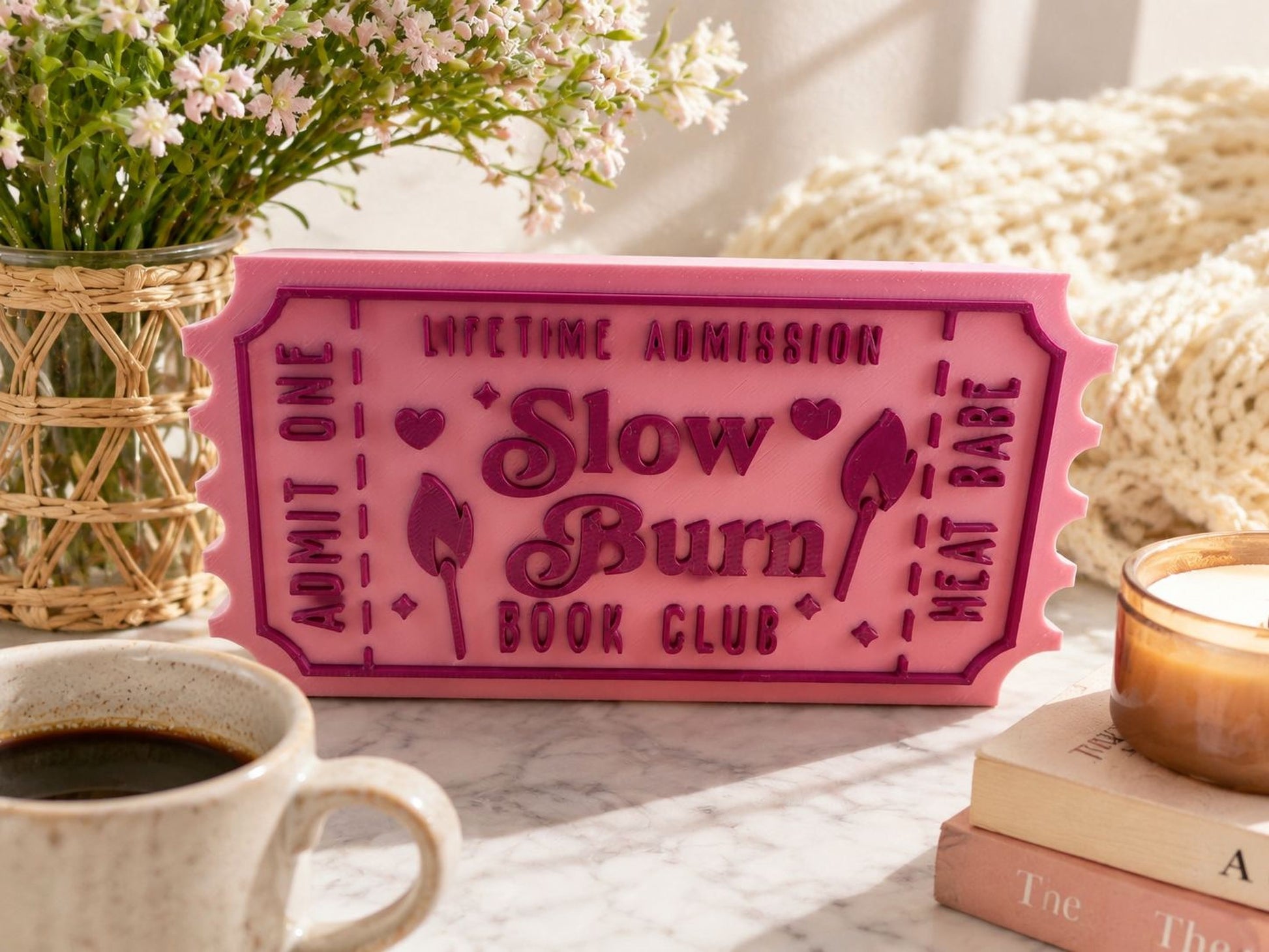 a pink ticket-shaped sign with the text "Lifetime Admission Slow Burn Book Club" on it, placed on a table next to a mug of coffee and a stack of books.