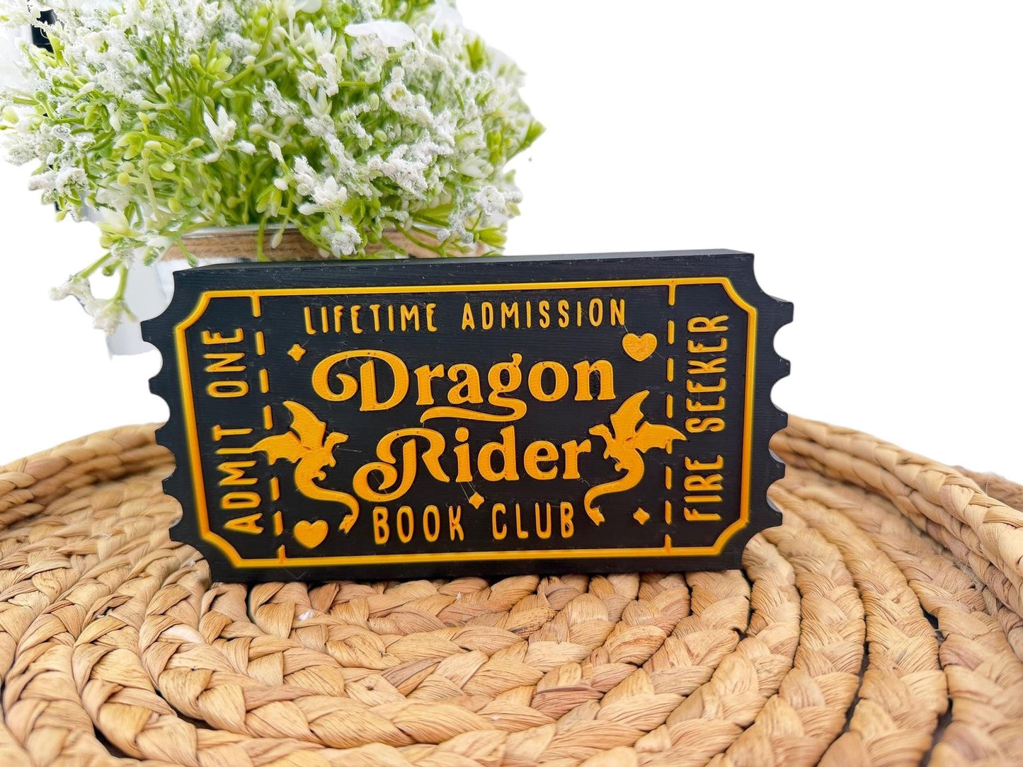A black and gold ticket-shaped sign with the text "Dragon Rider Book Club" printed on it, placed on a woven mat.