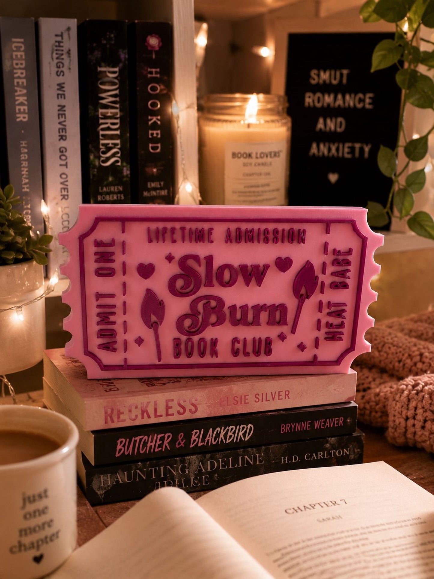 a pink ticket-shaped object with the text "Lifetime Admission Slow Burn Book Club" on it, surrounded by books, a candle, and a mug.