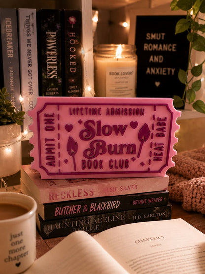 a pink ticket-shaped object with the text "Lifetime Admission Slow Burn Book Club" on it, surrounded by books, a candle, and a mug.