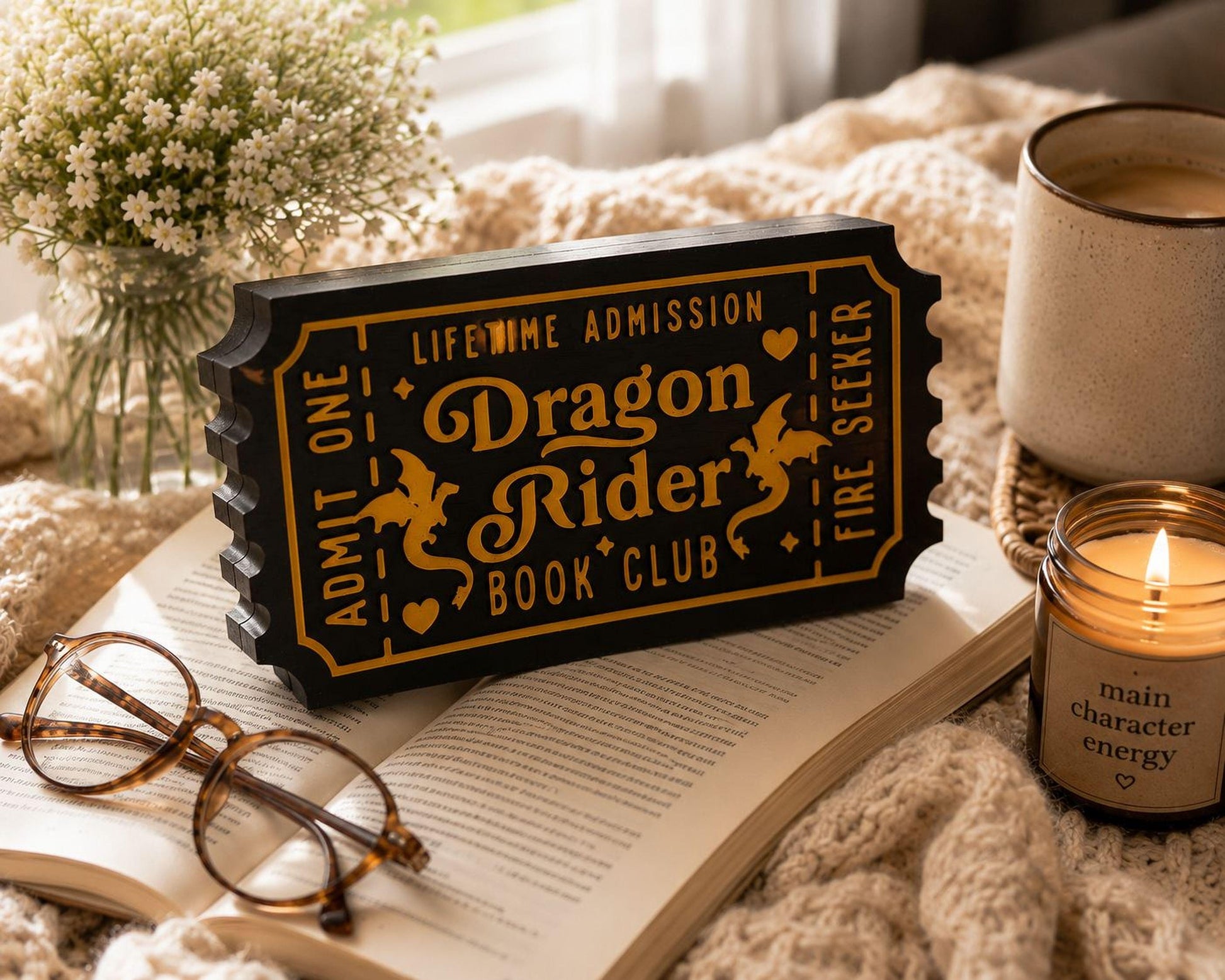 a black and gold ticket-shaped sign with the text "Dragon Rider Book Club" on it, placed on top of an open book. Next to the ticket-shaped sign is a pair of glasses, and a candle is visible in the background.
