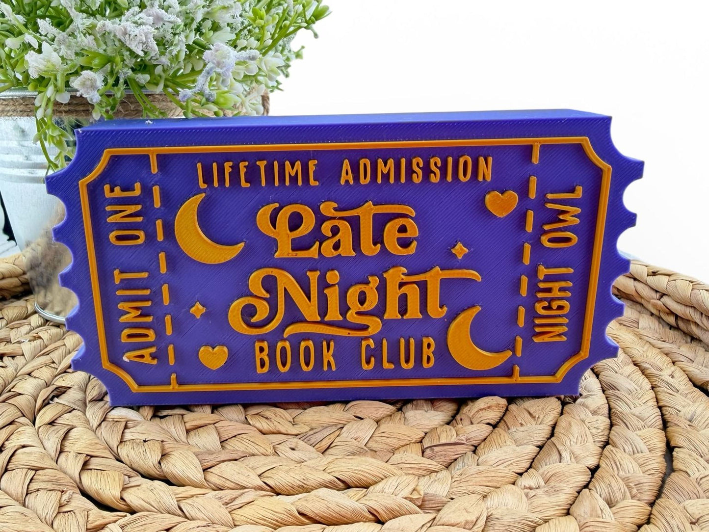 A purple ticket-shaped object with the text "Lifetime Admission Late Night Book Club" printed on it, placed on a woven mat.