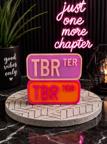 a pink and orange rectangular sign with the text "TBR" on it, placed on a black and white checkered surface. In the background, there is a black plant and a black and white geometric patterned plate.